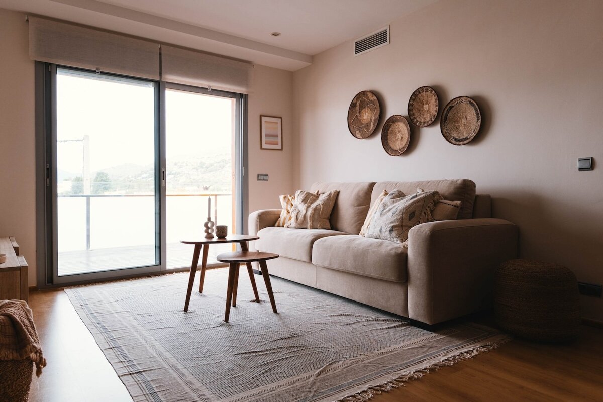 A cozy, neutral-toned living room with a beige sofa, patterned rug, wooden side tables, and wall baskets, opening to a bright outdoor view.