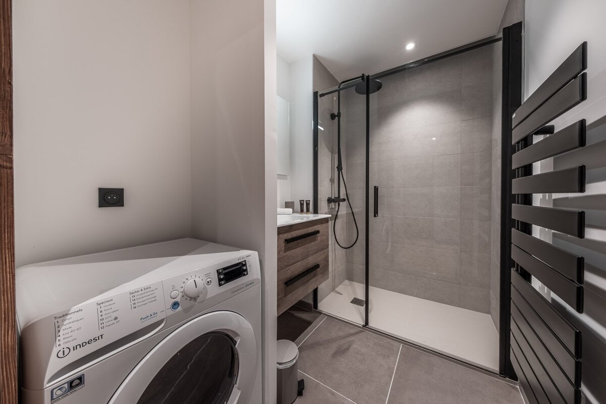 Modern bathroom featuring a white Indesit washing machine, a sleek glass-enclosed shower with black fixtures, and a wooden vanity with a sink.