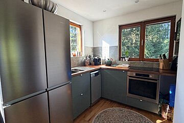 Modern kitchen with a large stainless steel fridge, sage green cabinets, wooden countertops, and two windows offering a lush tree view.