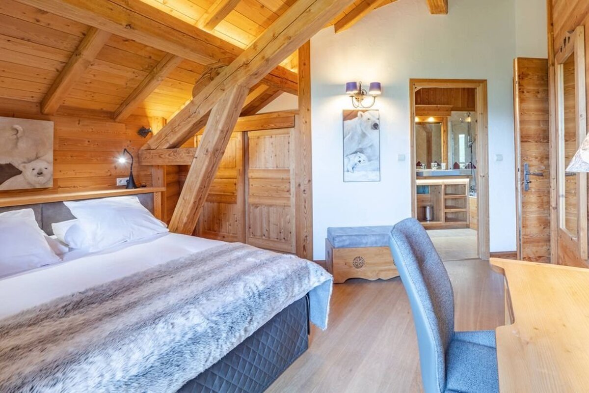 A cozy wooden chalet bedroom featuring vaulted ceilings, a bed with a faux-fur throw, and an open doorway leading to the bathroom.