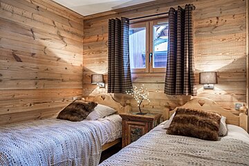 A cozy, wood-paneled chalet bedroom with two single beds, each with a furry pillow. A window with checked curtains is between the beds, above a rustic nightstand.