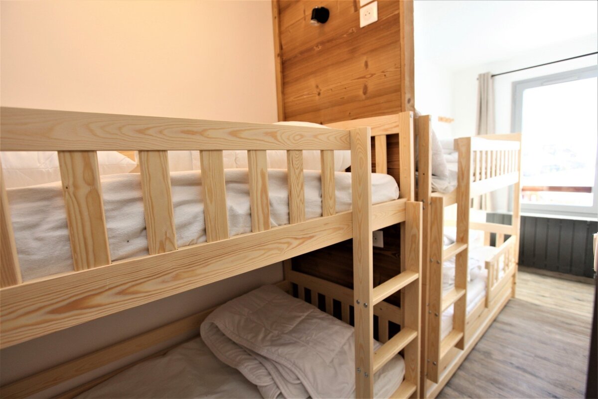 Light wooden bunk beds with white bedding fill a room, featuring wood-paneled walls and a window in the background.