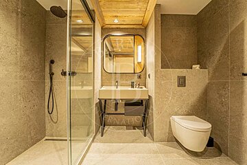 Sleek modern bathroom with grey stone tiles, wooden ceiling, walk-in glass shower, stylish sink with a rounded mirror, and a wall-mounted toilet.