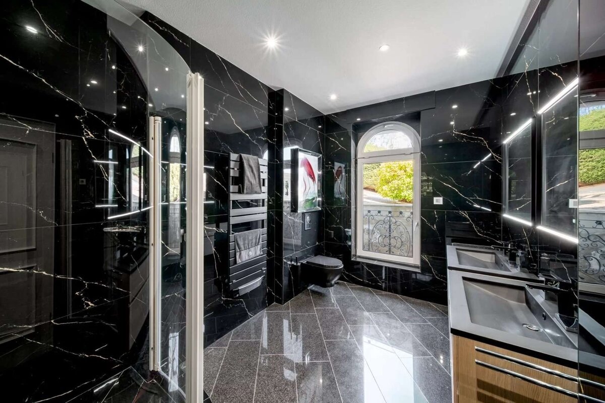 A sleek, modern bathroom with glossy black marble walls and floor, a double vanity, toilet, shower, and an arched window.