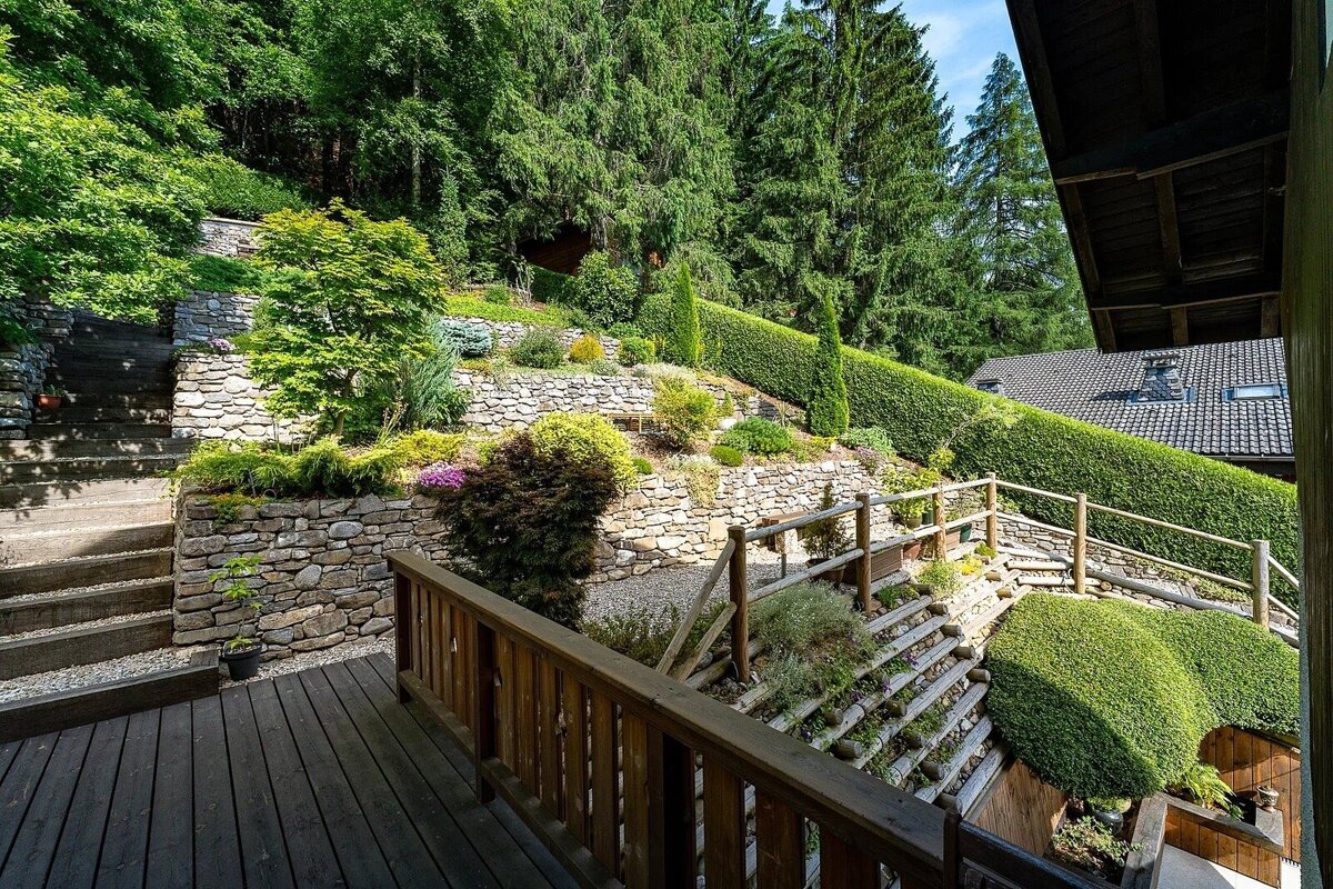 A scenic terraced garden with stone walls, wooden stairs, and vibrant greenery on a hillside, with a house roof in the background.