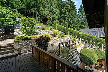 Lush terraced garden on a hillside, featuring stone walls, wooden steps, diverse greenery, hedges, and towering trees, with a house roof peeking through.