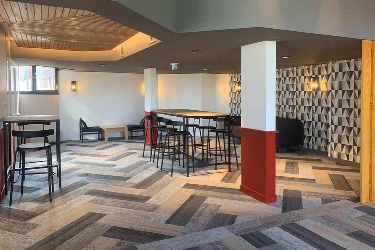 A modern communal lounge with herringbone flooring, high-top tables, bar stools, geometric wallpaper, and warm wooden ceiling details.
