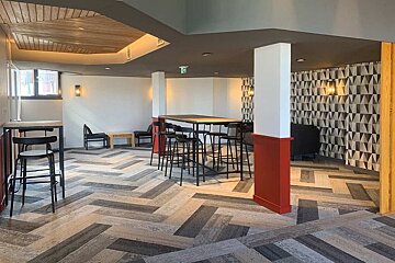 A modern communal lounge with herringbone flooring, high-top tables, bar stools, geometric wallpaper, and warm wooden ceiling details.