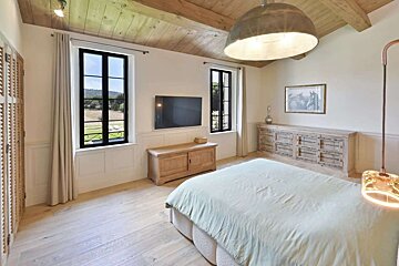 Spacious, bright bedroom with vaulted wood ceilings, large windows revealing nature, a light green bed, and rustic wooden furniture.
