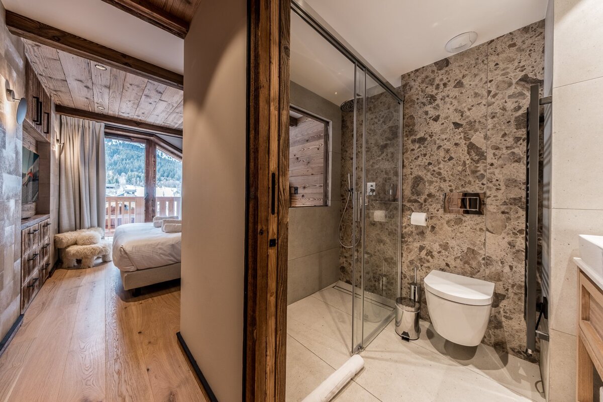A cozy chalet bedroom with a mountain view balcony flows into a modern bathroom featuring a glass shower, toilet, and stone-tiled walls.