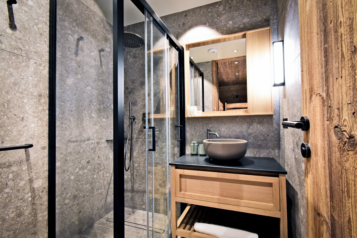 Modern bathroom featuring gray stone-tile walls, a glass shower, a light wood vanity with a bowl sink, and a rustic wooden door.