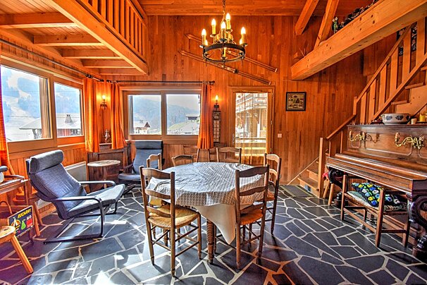 A rustic wooden chalet-style living/dining room with a stone floor, mountain views, a dining table, armchairs, and a piano.
