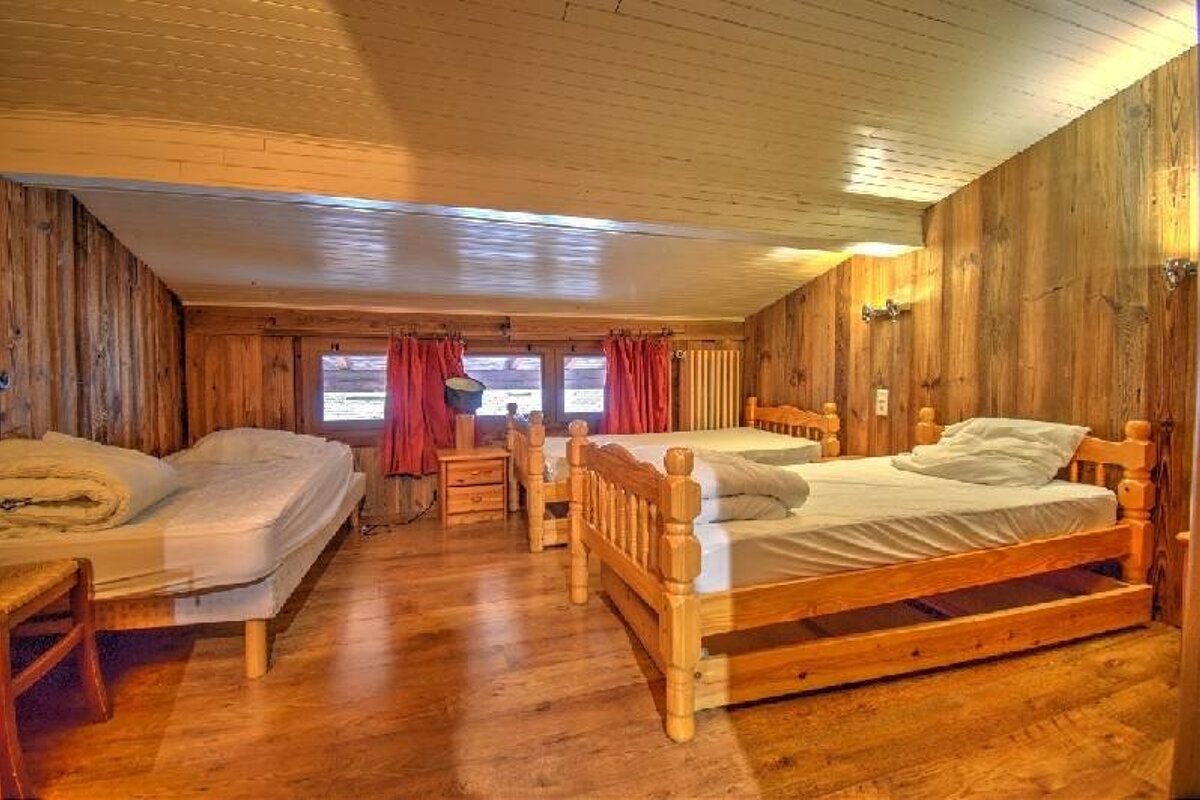 A rustic wooden attic bedroom with five single beds, white bedding, and red curtains on windows, giving a cozy, cabin-like feel.
