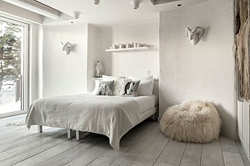 A cozy, white, arctic-themed bedroom featuring textured wavy walls, a bed with animal pillows, a furry beanbag, and a snowy outdoor view.