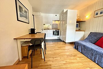 A small open-plan living space with a wooden dining table, black chairs, a blue sofa, and a compact white kitchen in the background.