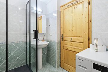 A compact bathroom featuring a glass shower, a pedestal sink, green tiled walls, a wooden door, and a Miele washing machine.