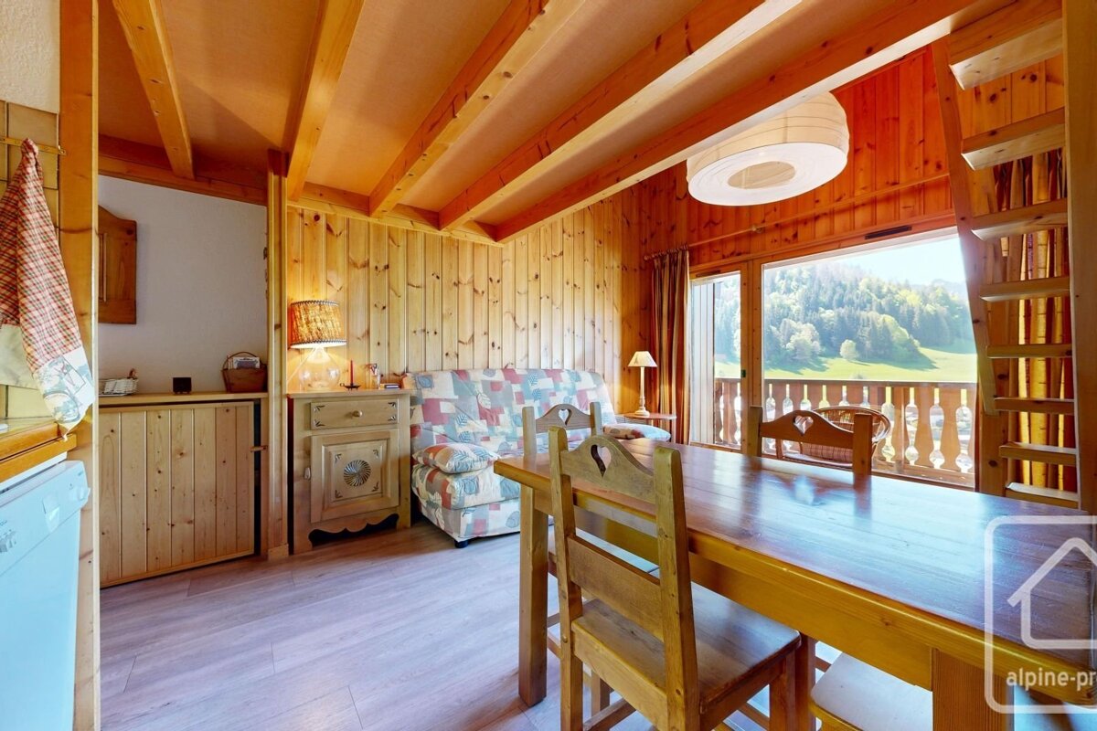Rustic wood-paneled room with dining set, patterned sofa, and balcony overlooking lush green mountains. Cozy chalet decor.