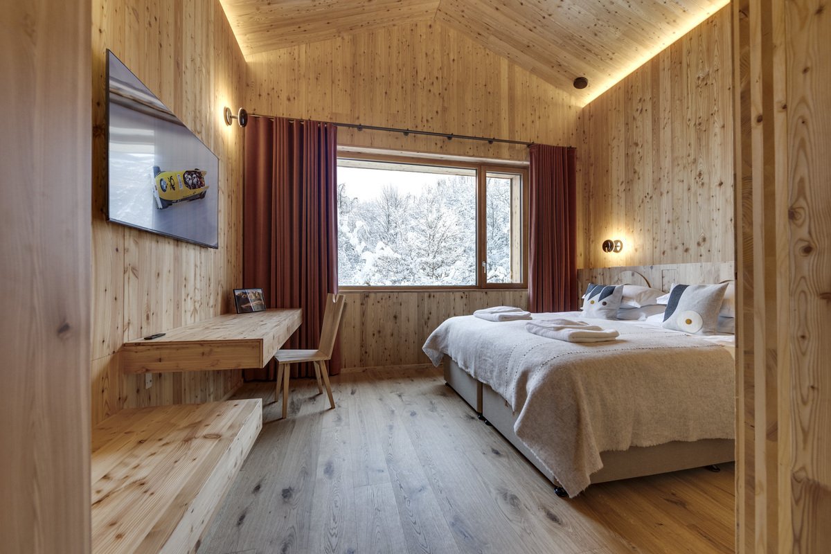 A cozy, wood-paneled bedroom featuring a comfortable bed, a floating desk, and a large window with a snowy forest view.