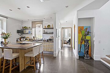 A modern, open-plan kitchen with a large island and bar stools, extending into a hallway featuring vibrant artwork and a glimpse of a room with a piano.