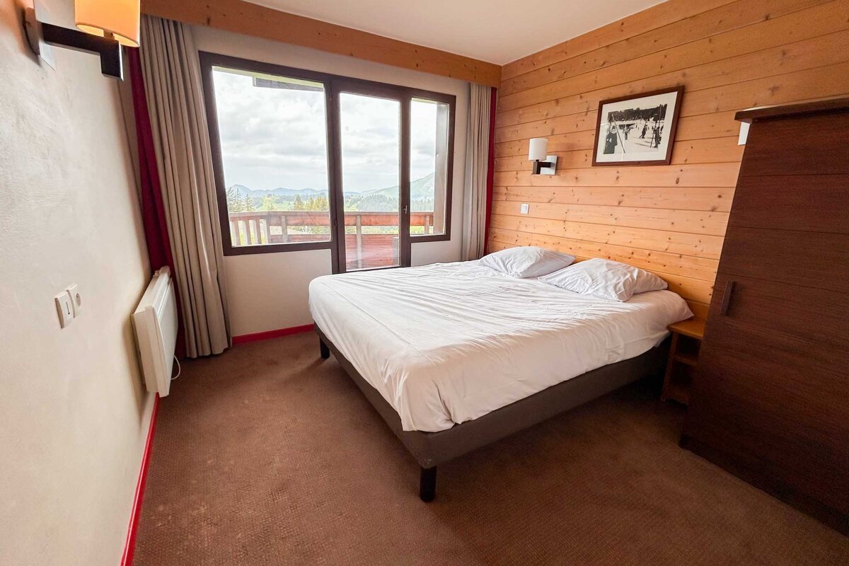 Cozy bedroom with a double bed, wood-paneled walls, and a large window offering a scenic mountain view from a balcony.