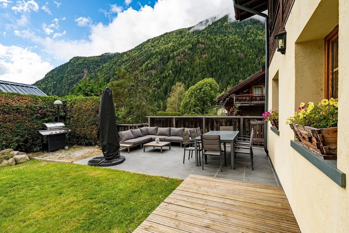 A spacious outdoor patio with a sofa, dining set, and BBQ, overlooking a lush green lawn and a forest-covered mountain range under a cloudy sky.