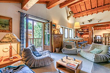 Cozy rustic chalet living room with exposed wooden beams, comfortable modern lounge chairs, and doors leading to a balcony.