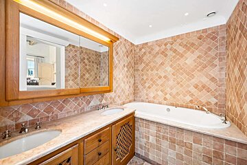 A tiled bathroom featuring a double-sink wooden vanity, a large mirror reflecting a bedroom, and a built-in bathtub.