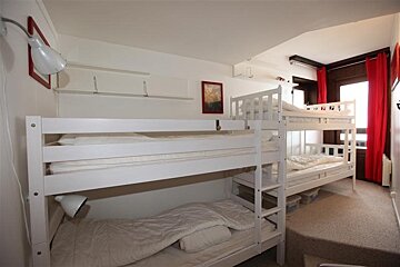 A bright room with two white bunk beds, white bedding, and red window curtains. White walls and simple decor.