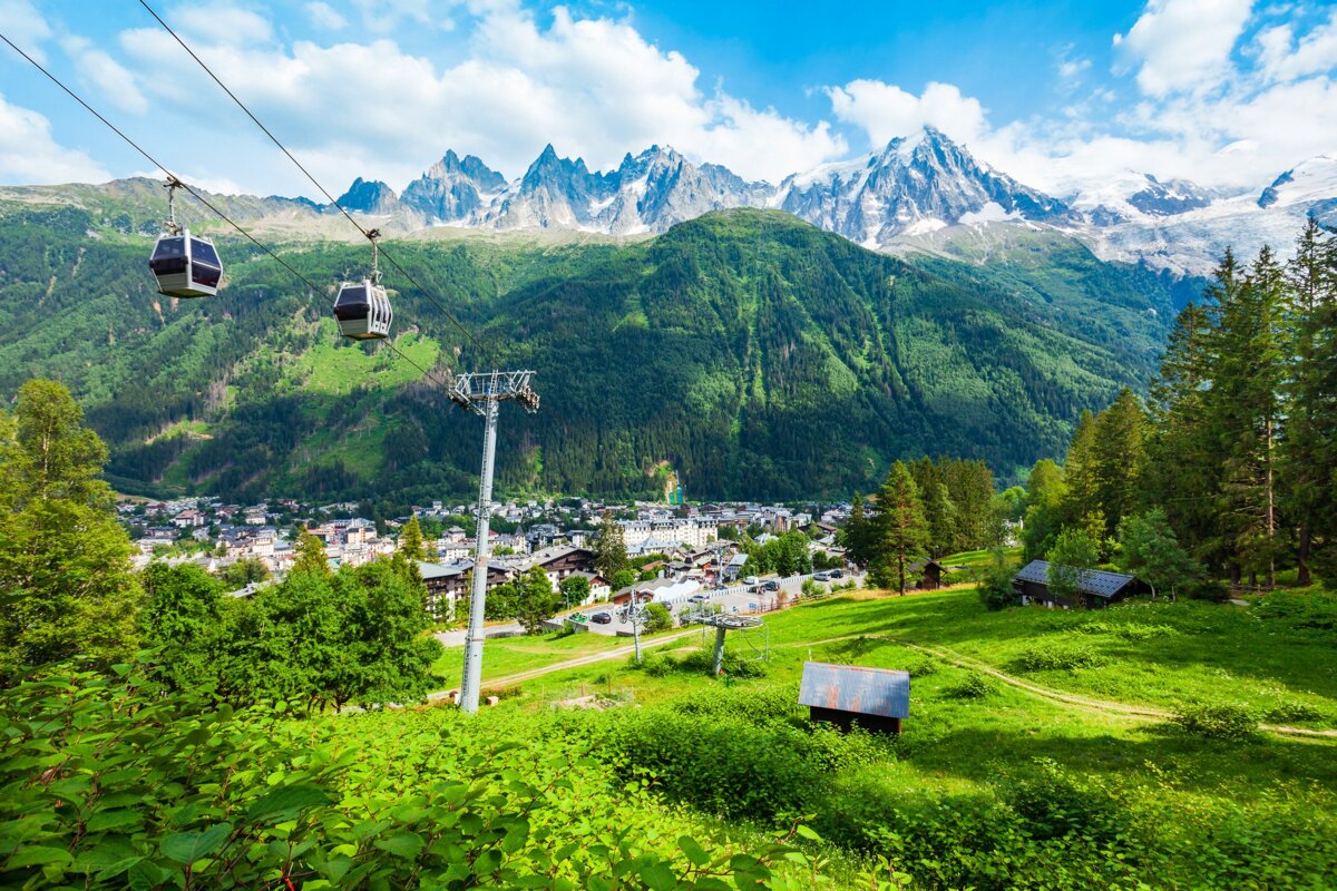 Gondolas ascend over a vibrant green valley, with a mountain town nestled below dramatic snow-capped peaks under a bright blue sky.