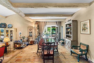 Spacious, rustic dining room with a long table, tiled floor, exposed beams, and French doors opening to a garden, decorated with traditional furniture.