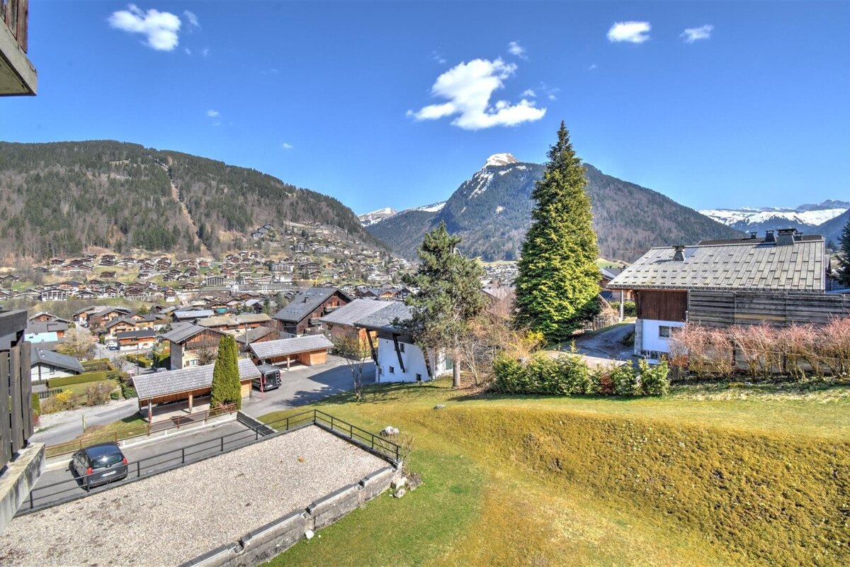 Alpine village with wooden chalets, green pine trees, and mountains under a clear blue sky.