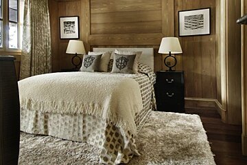 Rustic wood-paneled bedroom featuring a cozy bed, matching black nightstands with lamps, framed art, and a plush shag rug.