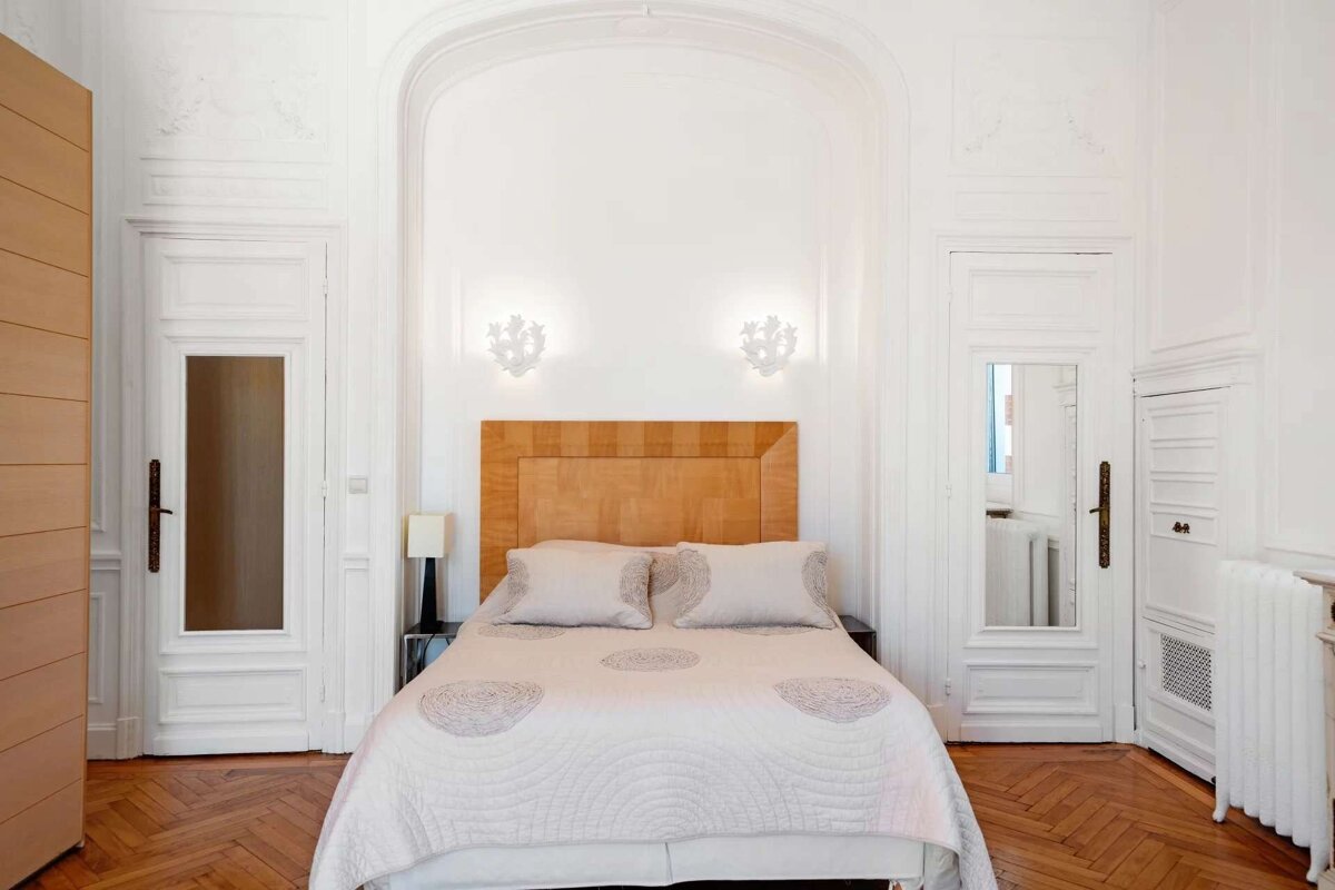 A bright, elegant bedroom features a wooden headboard bed flanked by ornate sconces within an arched alcove, set against white walls and a parquet floor.