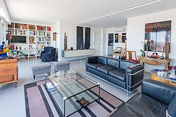 A bright, modern living room featuring black leather sofas, a glass coffee table on a striped rug, a wall-to-wall bookshelf, and contemporary art throughout.