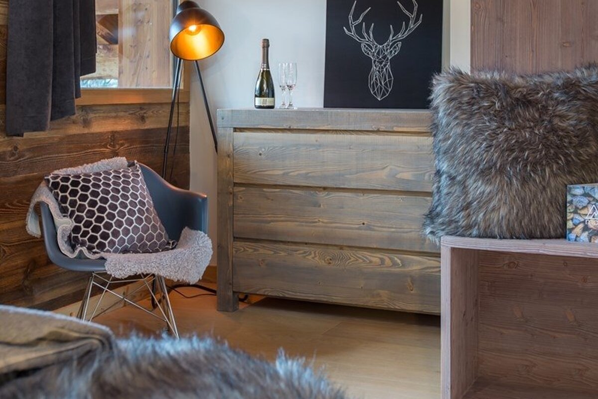 A cozy, rustic room featuring a wooden dresser, a modern armchair with fluffy throws, a floor lamp, and a deer art print.