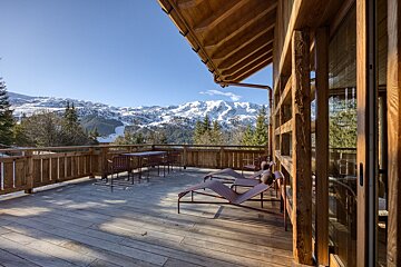 A sunny wooden chalet terrace with lounge chairs and a table, offering a breathtaking view of snow-covered mountains and pine forests.