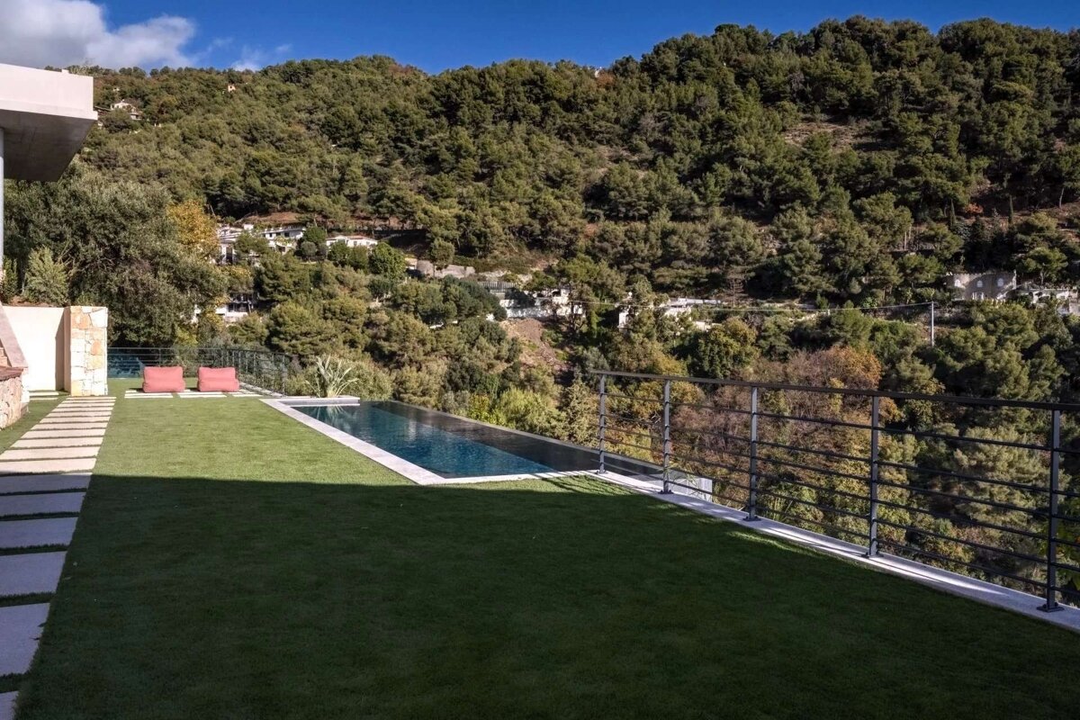 An infinity pool and green lawn overlook a densely forested mountain and distant homes under a clear blue sky.