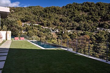 An infinity pool and green lawn overlook a densely forested mountain and distant homes under a clear blue sky.