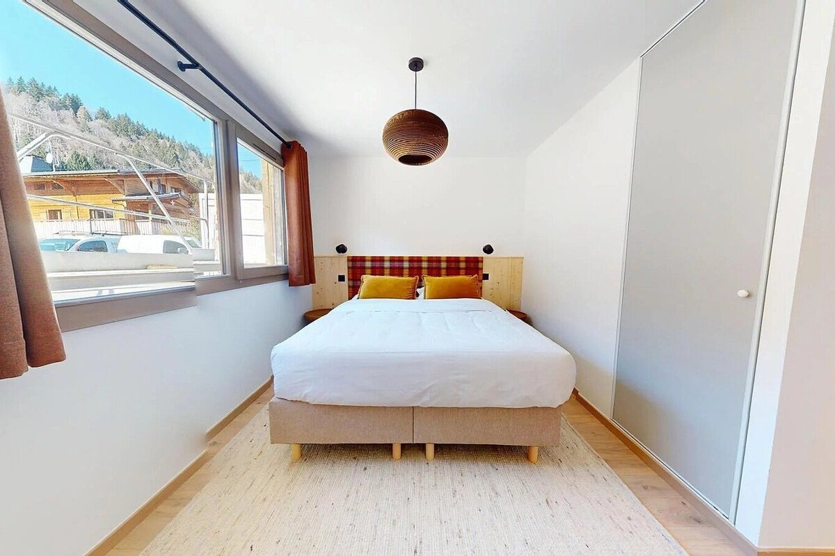 A bright bedroom with a large bed, plaid headboard, unique wooden ceiling light, and a wide window overlooking mountains and houses.