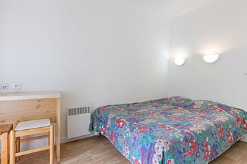 A simple bedroom with white walls, wooden floor, a bed with a colorful floral duvet, two wall lights, a wooden table with stools, and a heater.