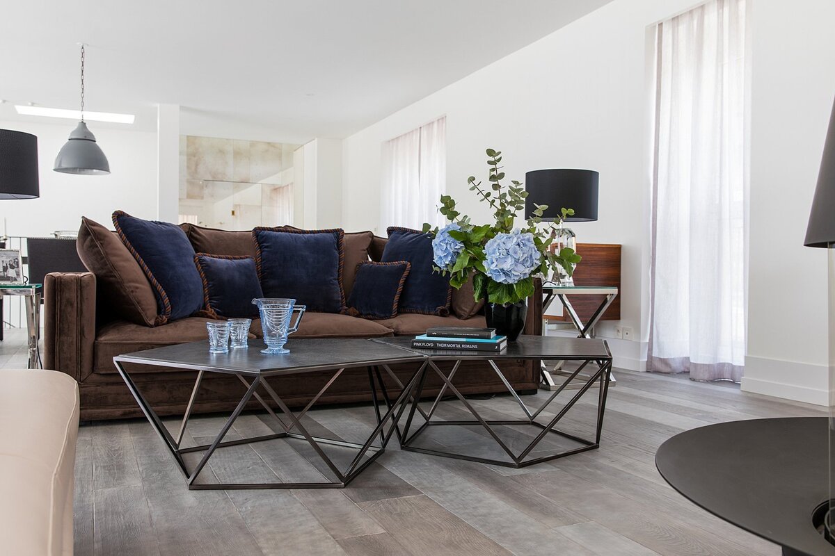 A modern living room features a brown velvet sofa with navy pillows, unique geometric coffee tables, and a vase of blue hydrangeas.