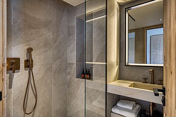 Modern bathroom with grey stone walls, a glass shower, bronze fixtures, and a vanity sink under a backlit mirror.