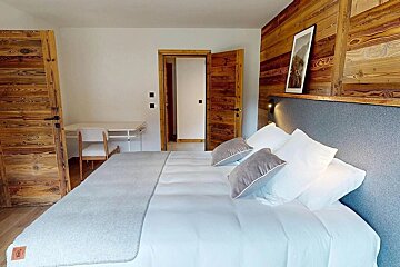 Modern bedroom with a white bed, grey accents, and rustic wooden doors and feature wall. A desk with a chair is visible.