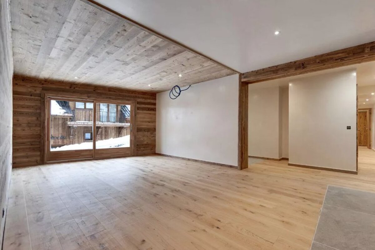 Empty modern chalet interior with light wood floor, rustic wood paneling, white walls, and a large window overlooking a snowy scene.