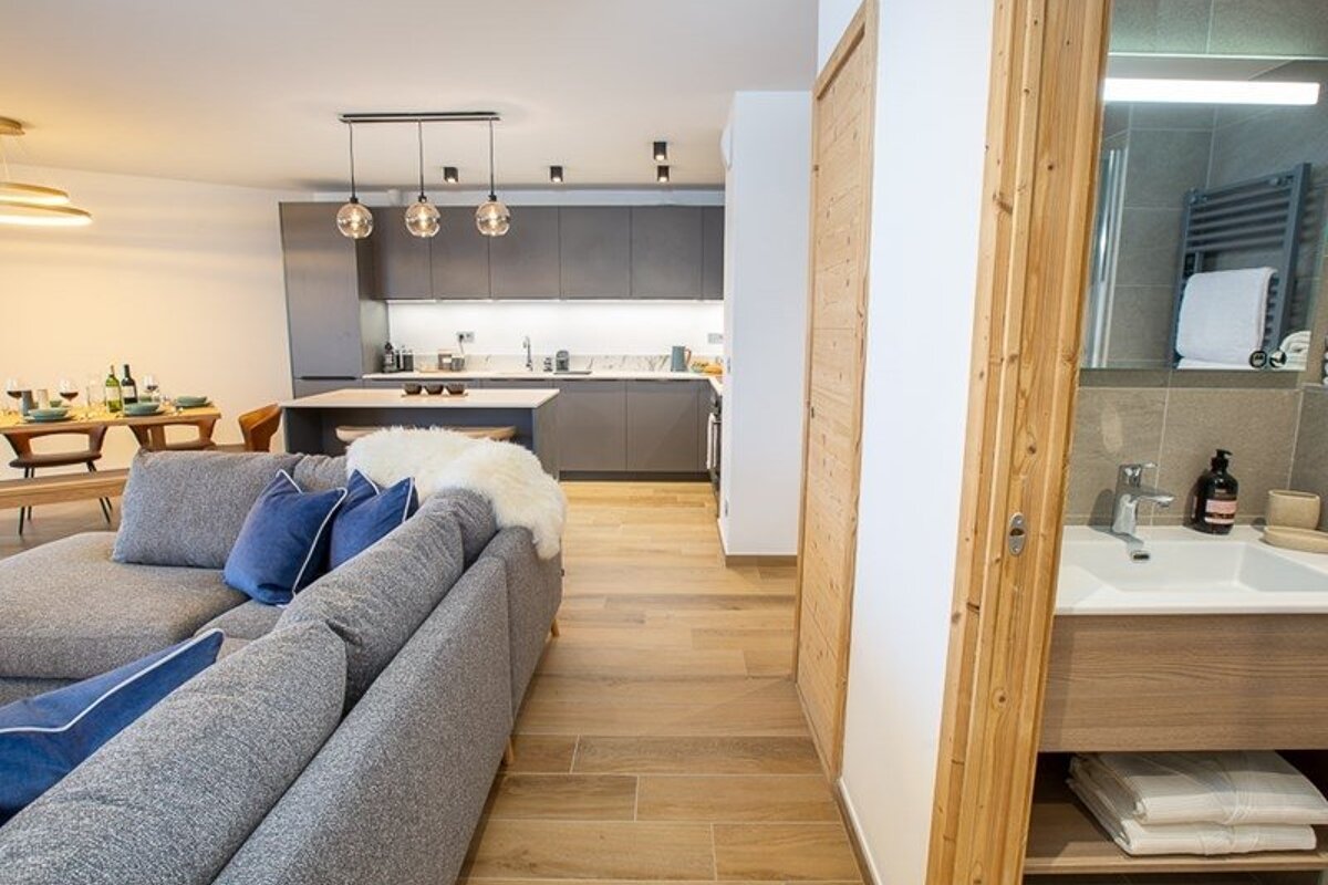 A modern open-plan apartment featuring a grey sofa, wooden flooring, a sleek kitchen, and a glimpse of the bathroom on the right.