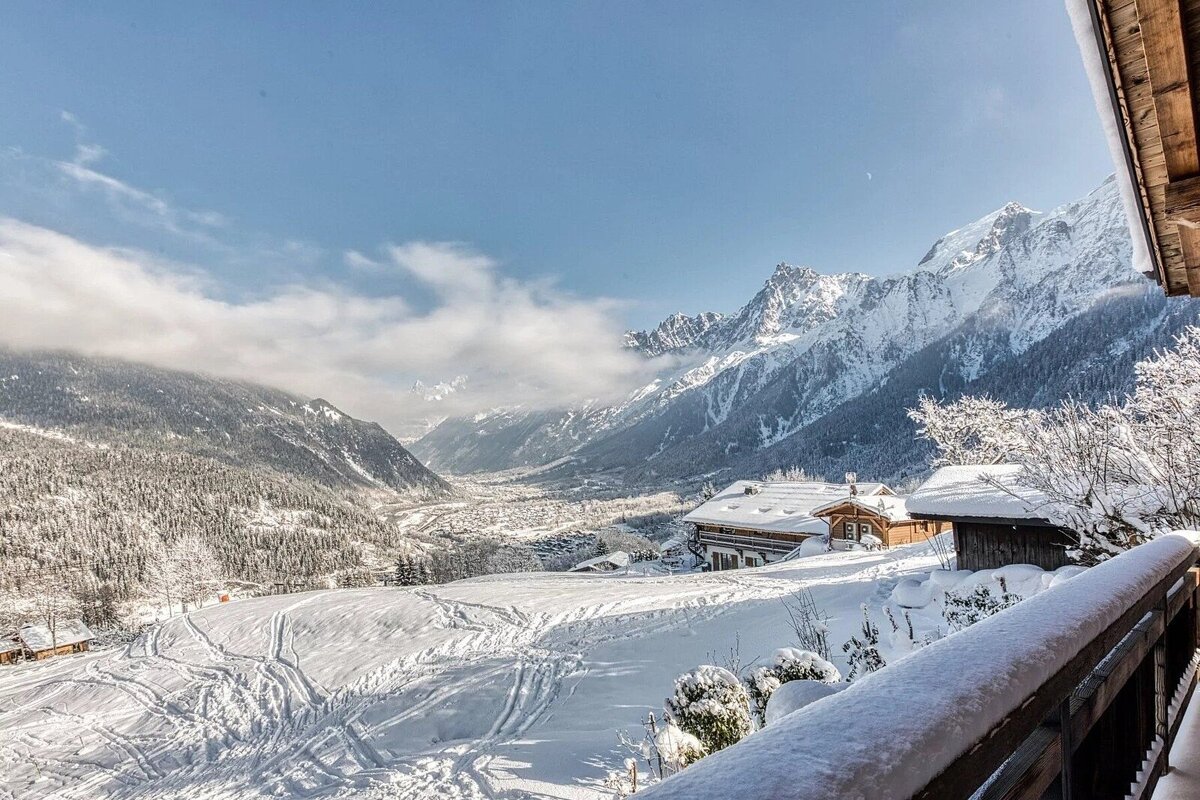 Panoramic view of a snowy mountain valley with chalets, ski tracks, and majestic, cloud-kissed peaks under a crisp blue sky.