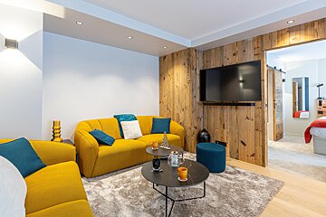 A modern living room with bright yellow sofas, a TV on a rustic wooden wall, coffee tables on a grey rug, opening to a bedroom.
