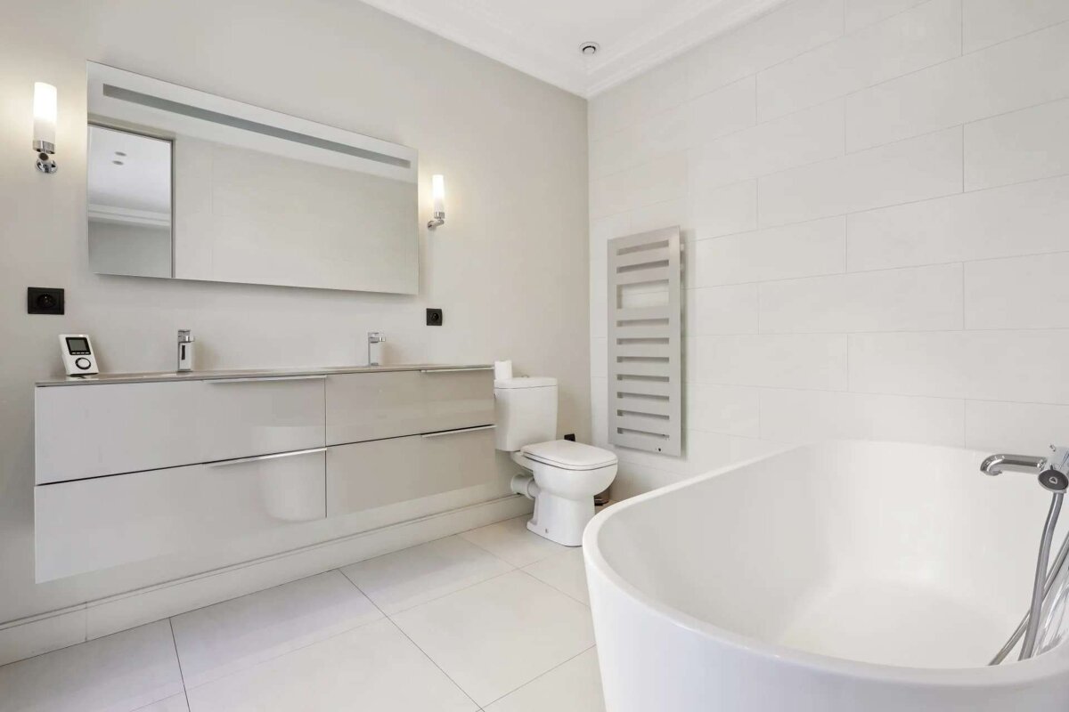 A modern, minimalist bathroom in white and light gray tones. Features a double vanity, large mirror, toilet, towel warmer, and freestanding tub.
