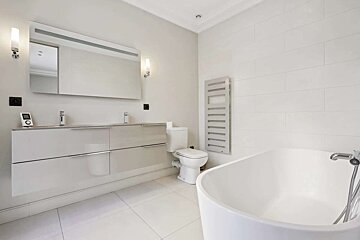 A modern, minimalist bathroom in white and light gray tones. Features a double vanity, large mirror, toilet, towel warmer, and freestanding tub.