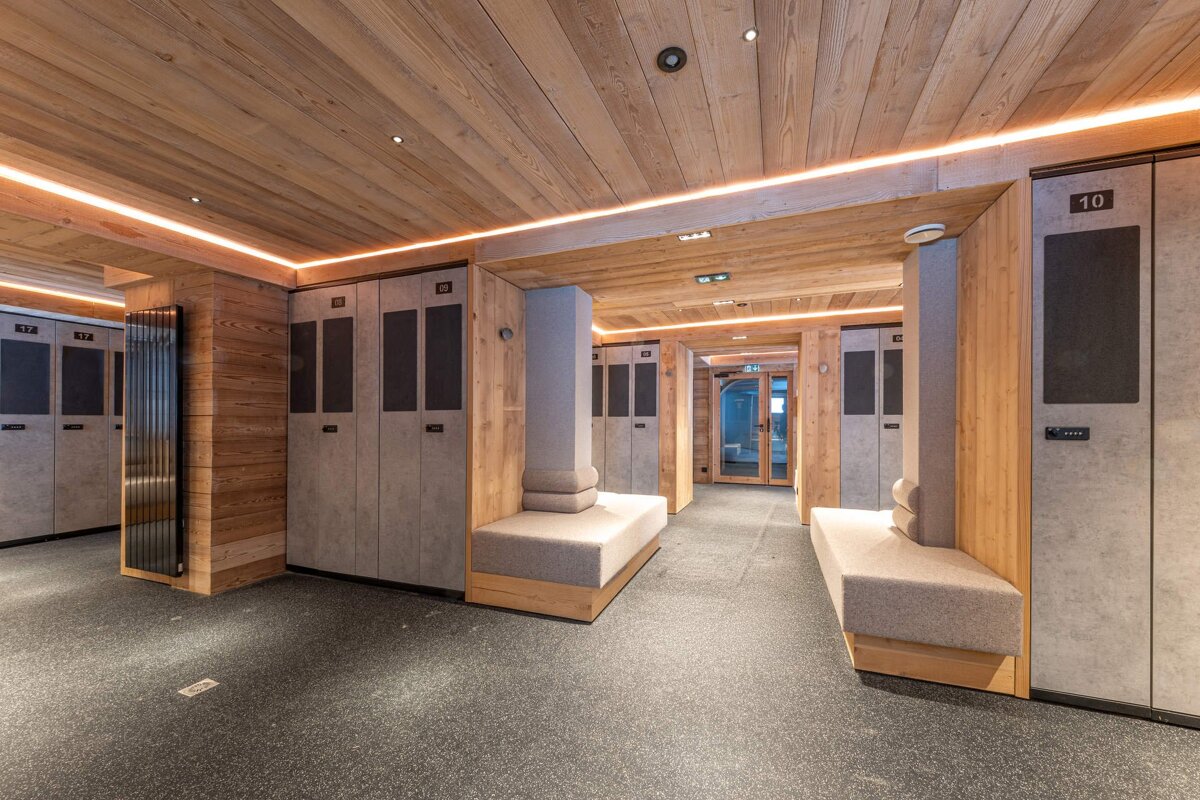 A wooden sauna with tiered benches, featuring a large window showcasing a vivid blue sky, snowy mountains over water, and an adjoining wooden room.
